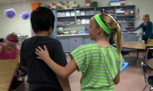 two children standing in a classroom. One child has a hand on the other child's back in comfort.