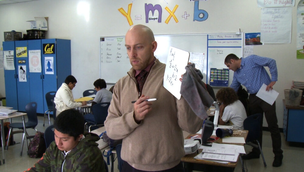 A male teacher standing in a classroom with seated students holding up a dry erase board with match equations written on it.